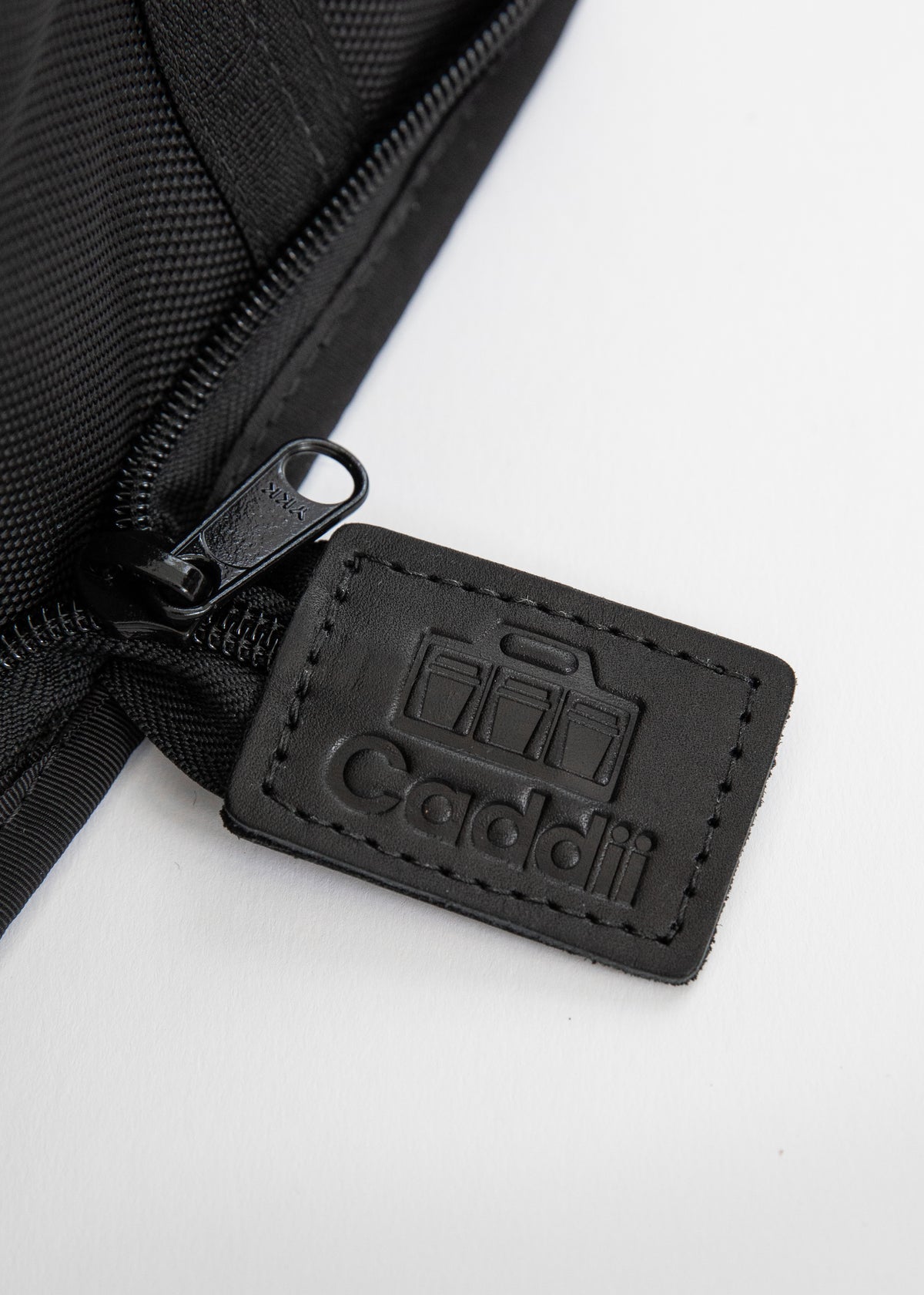 Close-up of a black zippered pouch with a stitched black leather tag embossed with the Caddii logo, on a white background.
