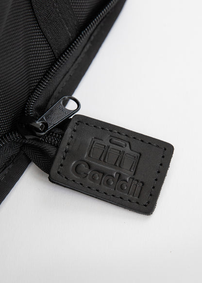 Close-up of a black zippered pouch with a stitched black leather tag embossed with the Caddii logo, on a white background.