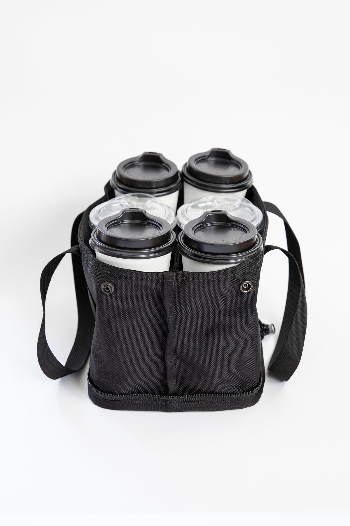 A black fabric four-cup coffee carrier bag holding four reusable travel mugs with lids, photographed on a clean white background.
